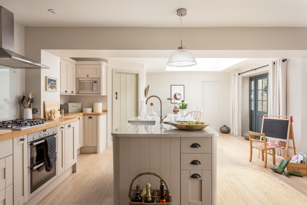Large country kitchen in Gloucestershire with recessed-panel cabinets, beige cabinets, wood benchtops, stainless steel appliances, with island, a farmhouse sink, beige splashback, glass sheet splashback, light hardwood floors and beige floor.