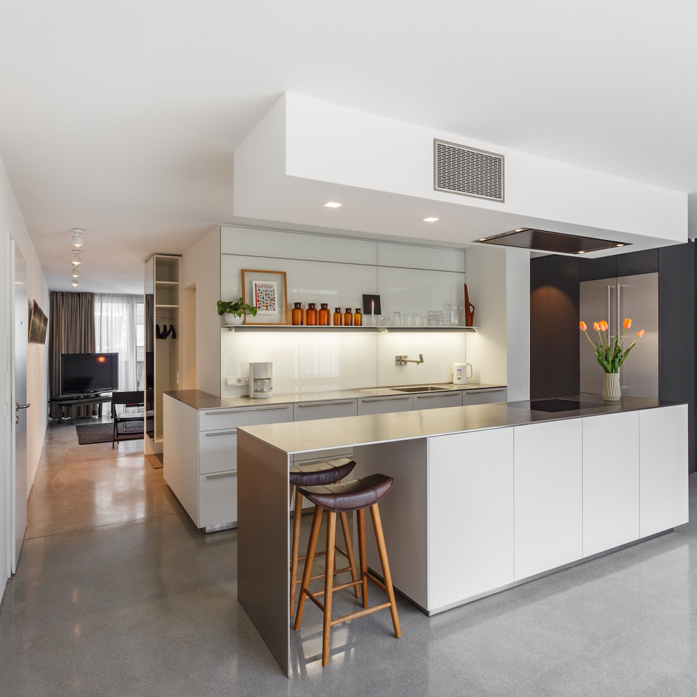 Photo of a mid-sized contemporary single-wall open plan kitchen in Hamburg with flat-panel cabinets, white cabinets, stainless steel benchtops, white splashback, glass sheet splashback, concrete floors, with island, grey floor and an integrated sink.