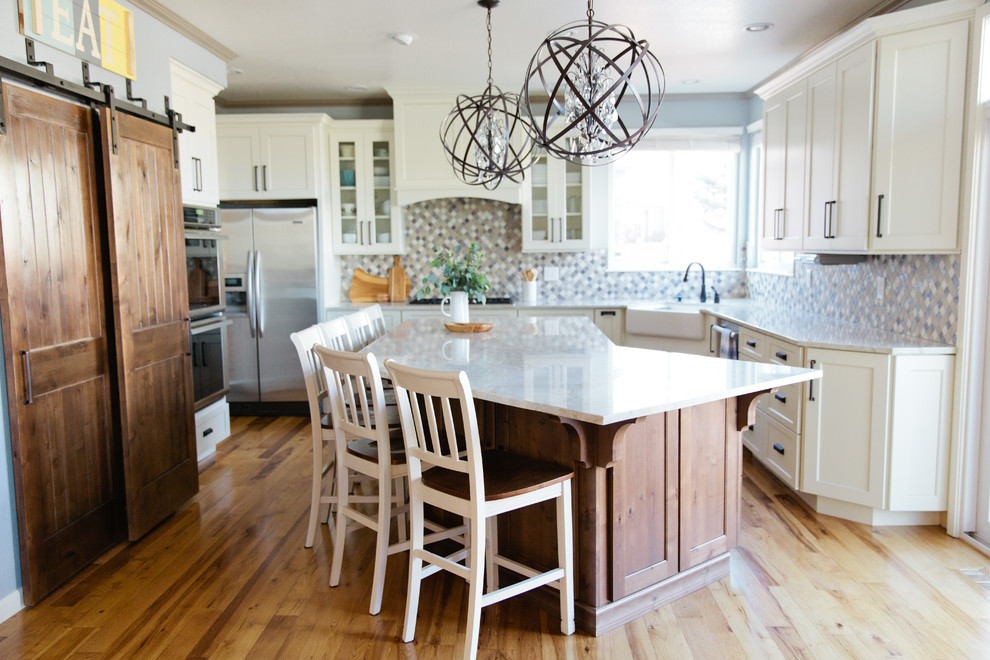 Inspiration for a large transitional u-shaped light wood floor eat-in kitchen remodel in Denver with a farmhouse sink, shaker cabinets, white cabinets, quartz countertops, multicolored backsplash, porcelain backsplash, stainless steel appliances and an island