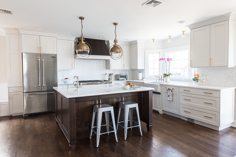This is an example of a mid-sized transitional l-shaped eat-in kitchen in New York with a farmhouse sink, white cabinets, white splashback, stainless steel appliances, with island, brown floor, flat-panel cabinets, quartz benchtops, ceramic splashback, medium hardwood floors and white benchtop.