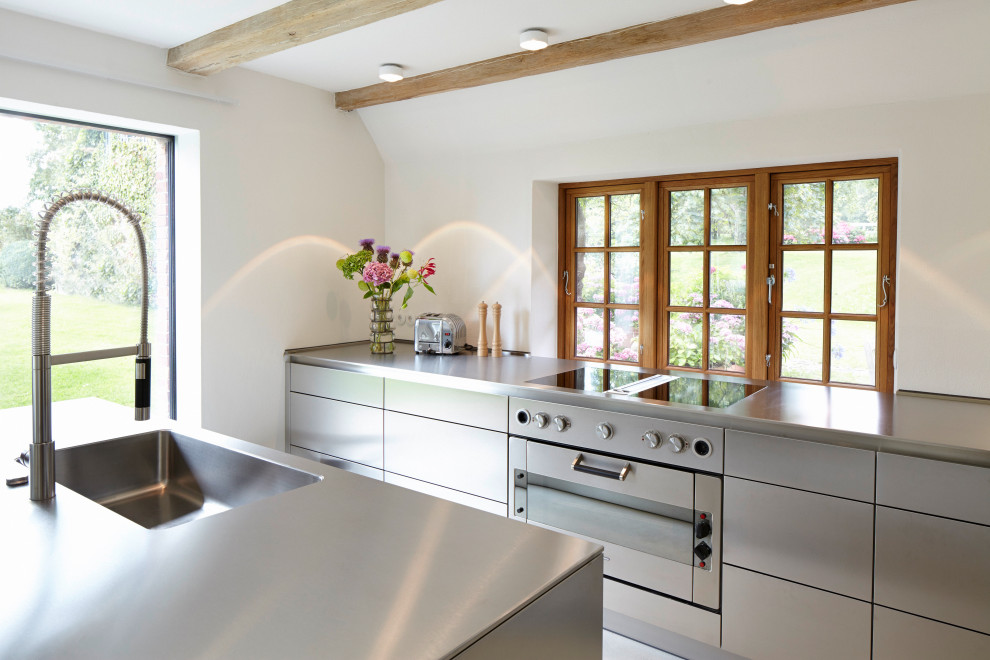 This is an example of a country galley kitchen in Hamburg with an integrated sink, flat-panel cabinets, grey cabinets, stainless steel benchtops, with island, grey floor, grey benchtop and exposed beam.