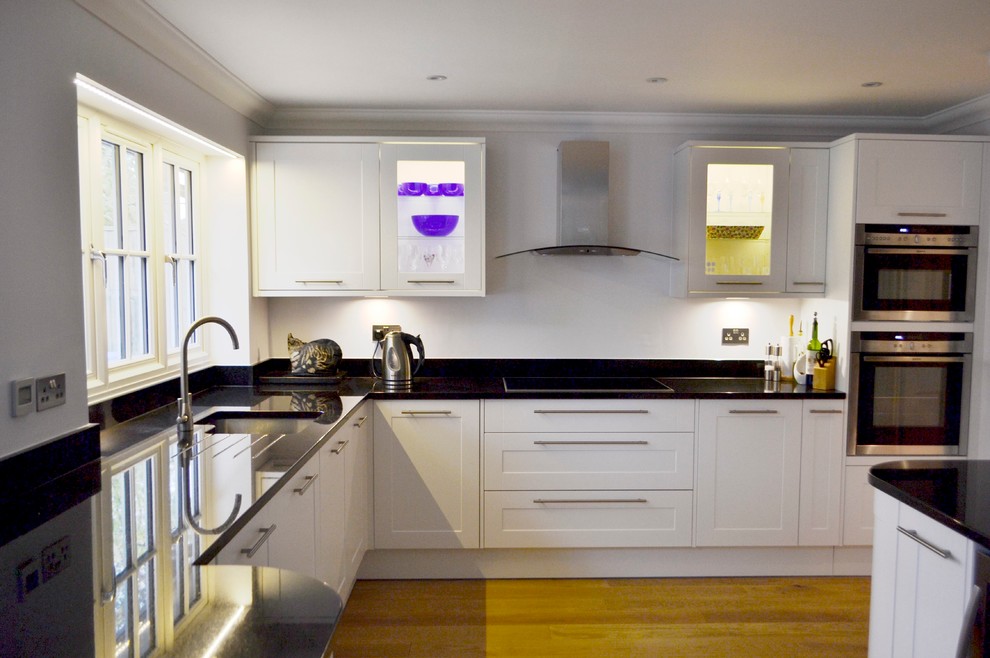 Example of a mid-sized trendy u-shaped light wood floor enclosed kitchen design in Surrey with a drop-in sink, shaker cabinets, white cabinets, granite countertops, black backsplash, stainless steel appliances and an island