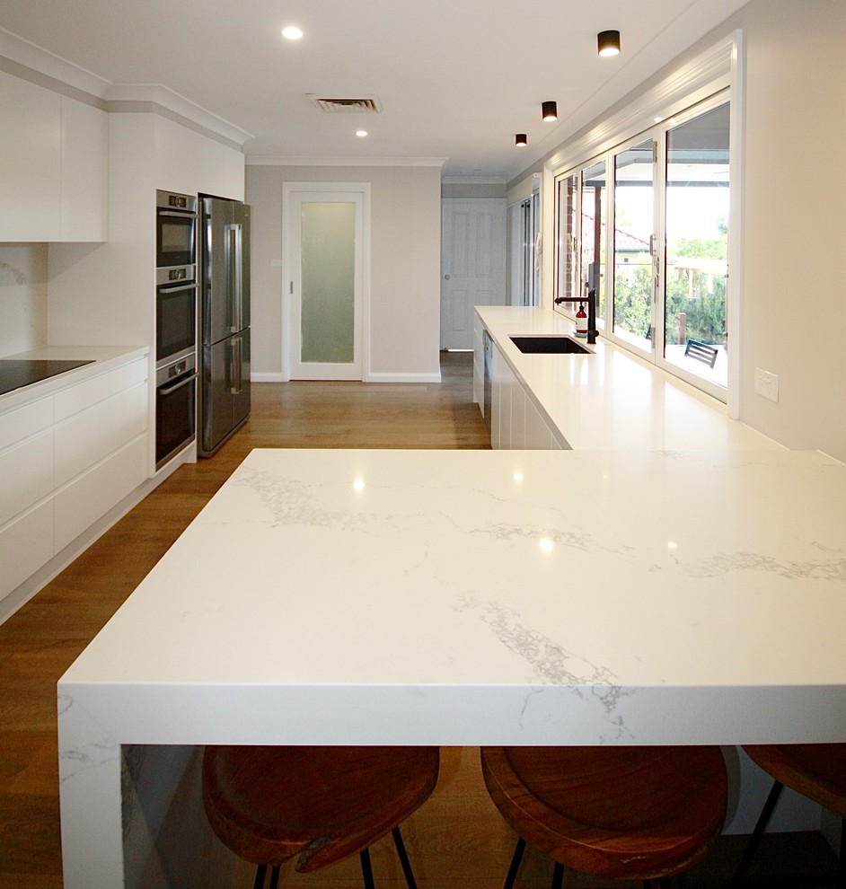 Mid-sized contemporary galley separate kitchen in Sydney with an undermount sink, flat-panel cabinets, white cabinets, quartz benchtops, white splashback, stone slab splashback, black appliances, light hardwood floors, with island and brown floor.