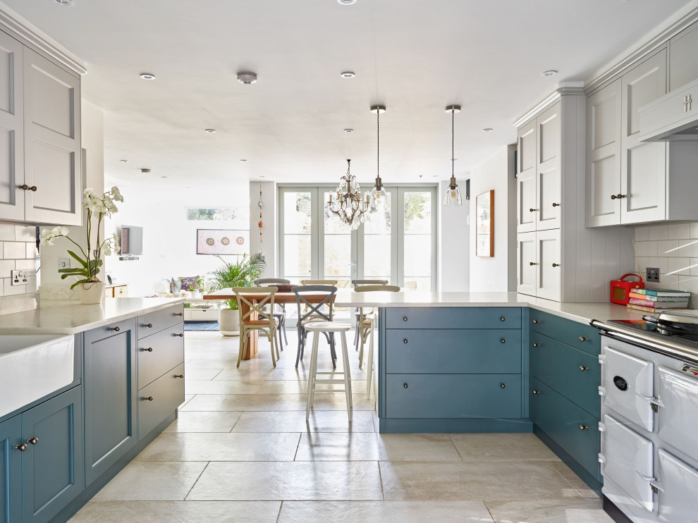 Traditional u-shaped separate kitchen in London with a farmhouse sink, recessed-panel cabinets, blue cabinets, white splashback, subway tile splashback, stainless steel appliances, a peninsula, grey floor and white benchtop.