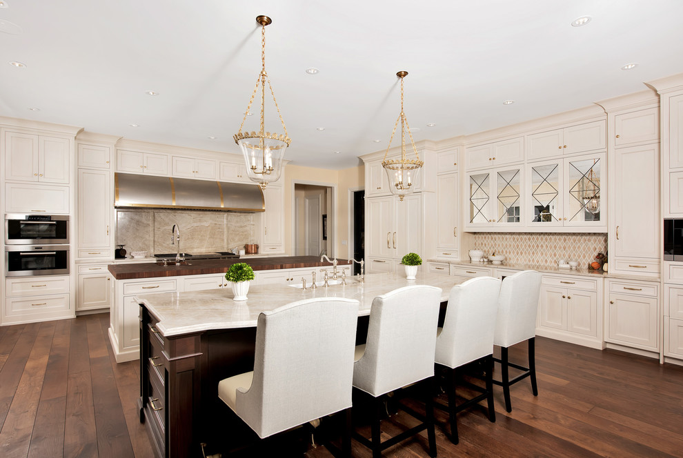 This is an example of an expansive traditional u-shaped eat-in kitchen in Chicago with shaker cabinets, beige cabinets, beige splashback, dark hardwood floors, multiple islands, an undermount sink, marble benchtops, stone slab splashback, stainless steel appliances and brown floor.
