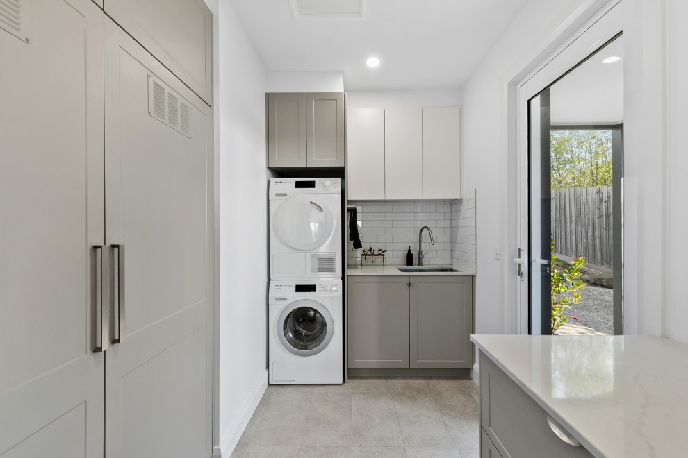 Design ideas for a mid-sized transitional dedicated laundry room in Geelong with an undermount sink, shaker cabinets, grey cabinets, quartz benchtops, white splashback, ceramic splashback, white walls, ceramic floors, a stacked washer and dryer and grey floor.