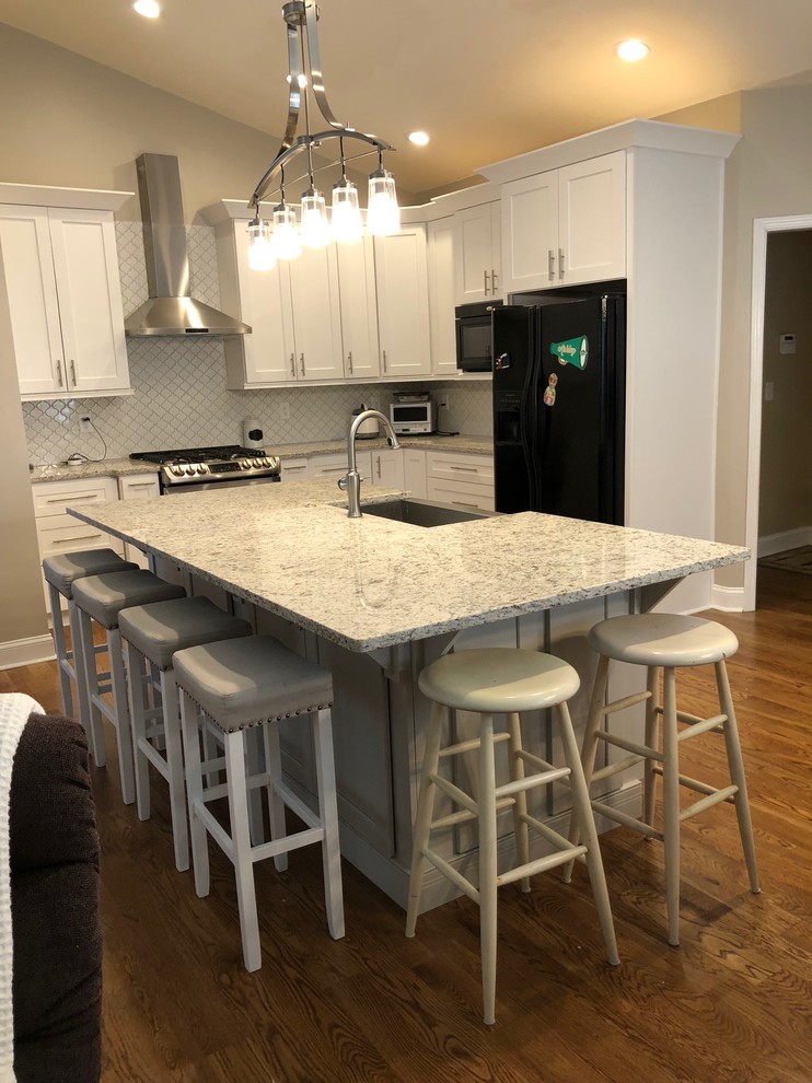 Mid-sized modern l-shaped eat-in kitchen in Atlanta with a farmhouse sink, shaker cabinets, white cabinets, granite benchtops, white splashback, ceramic splashback, stainless steel appliances, medium hardwood floors, with island and brown floor.