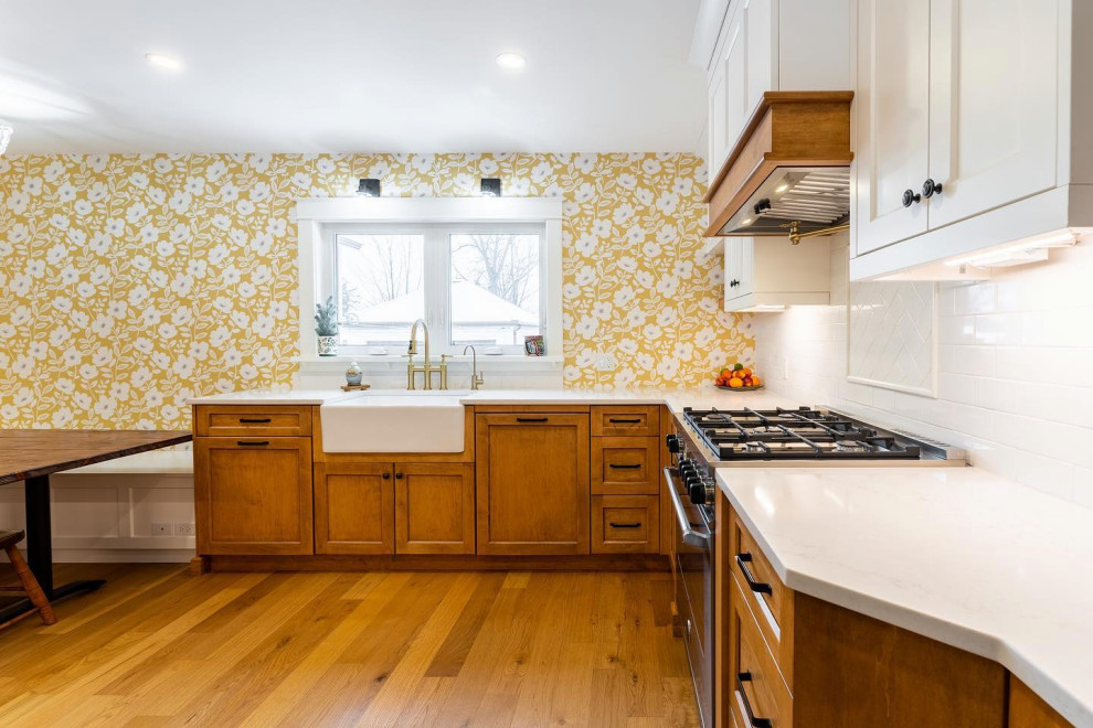 Photo of a mid-sized country l-shaped eat-in kitchen in Toronto with a farmhouse sink, shaker cabinets, brown cabinets, quartz benchtops, white splashback, stainless steel appliances, light hardwood floors, no island, orange floor, white benchtop and subway tile splashback.