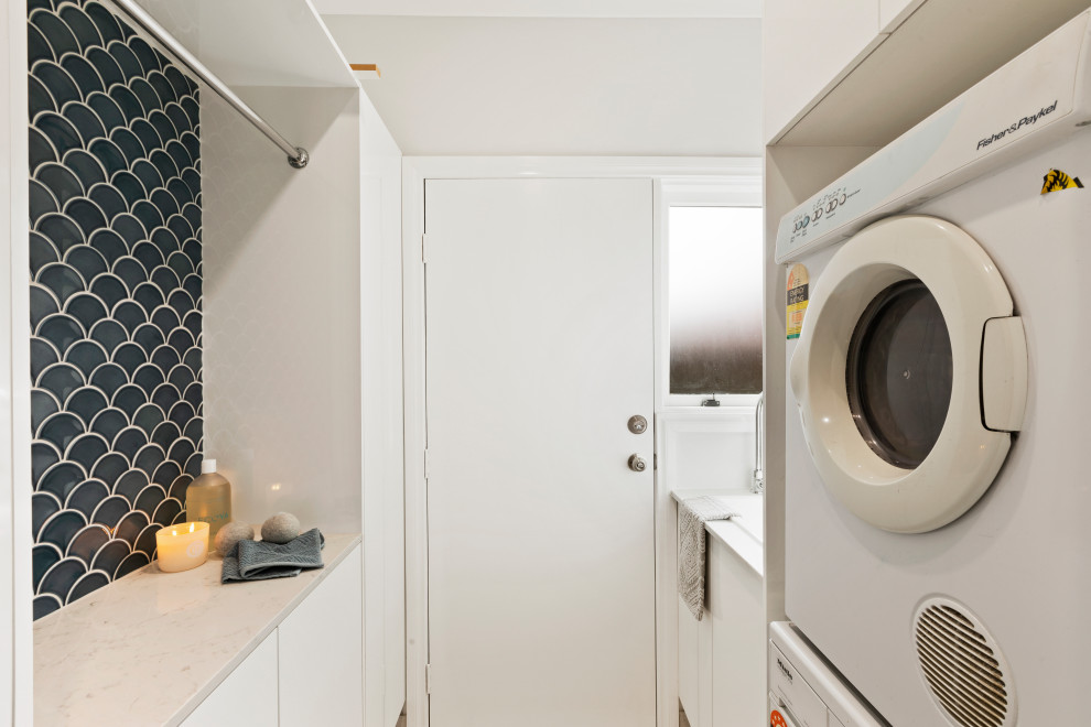 Small traditional galley dedicated laundry room in Sydney with a drop-in sink, flat-panel cabinets, blue splashback, mosaic tile splashback, white walls, a stacked washer and dryer and white benchtop.
