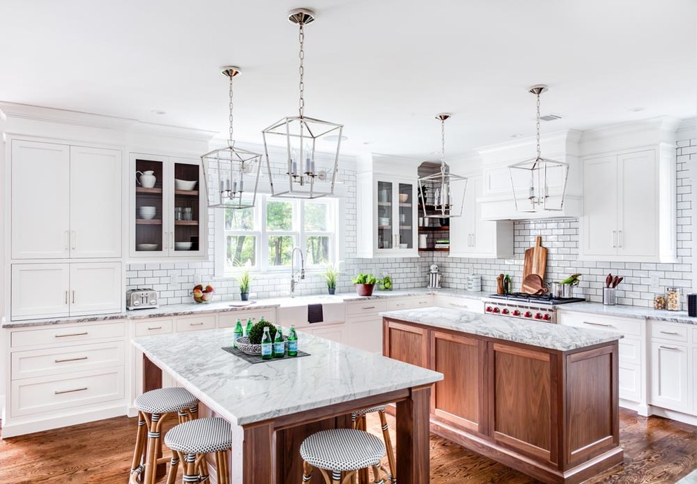 Mid-sized traditional l-shaped kitchen in Other with a farmhouse sink, white cabinets, marble benchtops, white splashback, subway tile splashback, stainless steel appliances, multiple islands, shaker cabinets and medium hardwood floors.