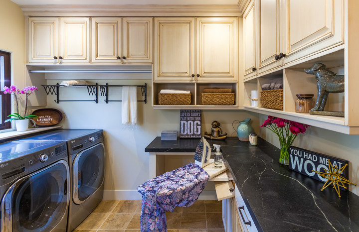 Photo of a large country u-shaped utility room in San Diego with raised-panel cabinets, soapstone benchtops, beige walls, travertine floors, a side-by-side washer and dryer, black benchtop and beige cabinets.