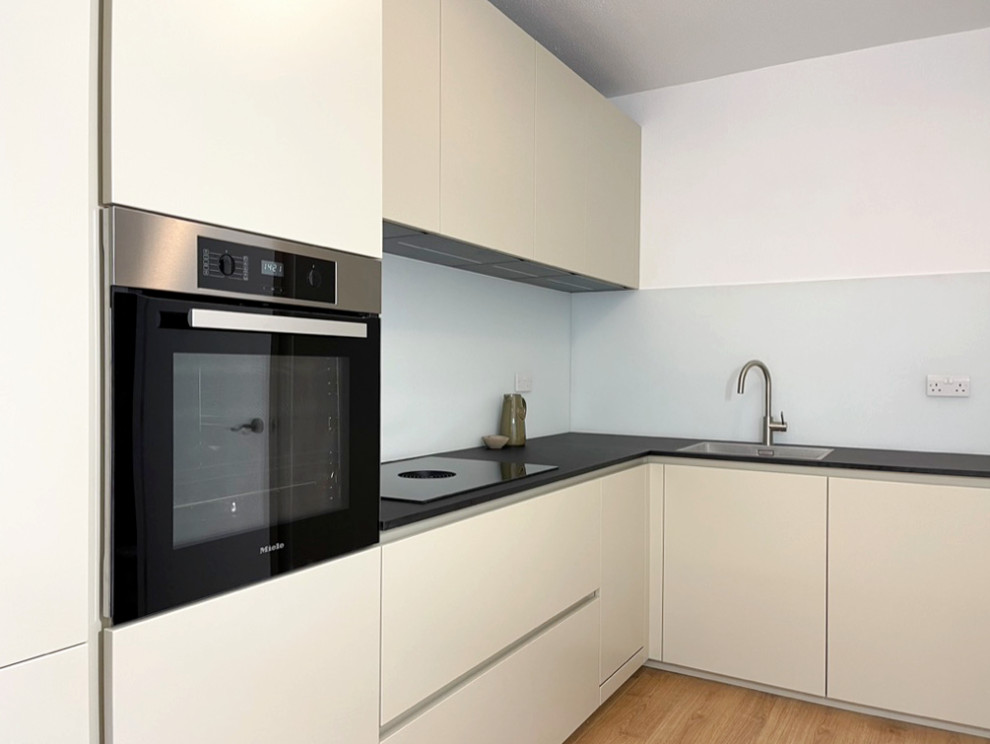 Inspiration for a small modern l-shaped enclosed kitchen in London with an integrated sink, flat-panel cabinets, beige cabinets, laminate countertops, white splashback, glass sheet splashback, black appliances, laminate floors, no island, brown floors and black worktops.