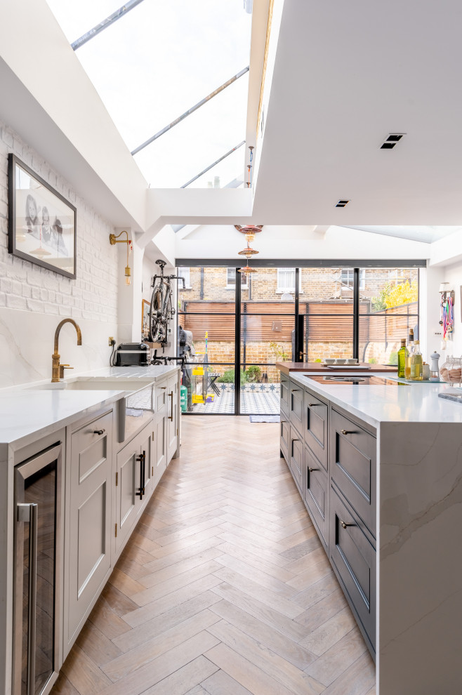 Inspiration for a large traditional galley open plan kitchen in London with a drop-in sink, recessed-panel cabinets, beige cabinets, marble benchtops, white splashback, marble splashback, black appliances, light hardwood floors, with island, brown floor, white benchtop and vaulted.