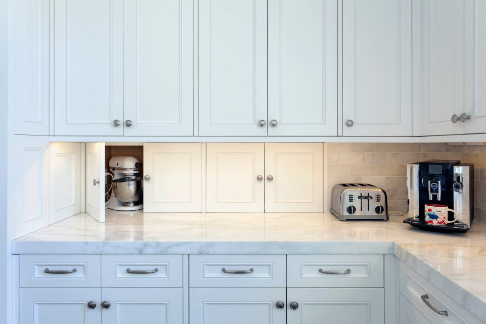 Inredning av ett klassiskt mycket stort vit vitt kök, med en undermonterad diskho, luckor med upphöjd panel, vita skåp, marmorbänkskiva, vitt stänkskydd, stänkskydd i marmor, rostfria vitvaror, mellanmörkt trägolv, en köksö och brunt golv
