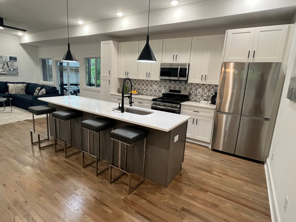 Photo of a mid-sized contemporary single-wall eat-in kitchen in Atlanta with a drop-in sink, recessed-panel cabinets, white cabinets, quartzite benchtops, grey splashback, ceramic splashback, stainless steel appliances, light hardwood floors, with island, brown floor and white benchtop.