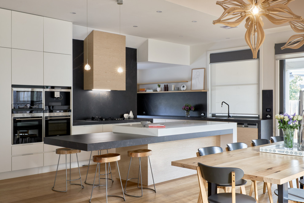 Design ideas for a contemporary u-shaped eat-in kitchen in Melbourne with an undermount sink, flat-panel cabinets, white cabinets, black splashback, panelled appliances, medium hardwood floors, with island, brown floor and black benchtop.