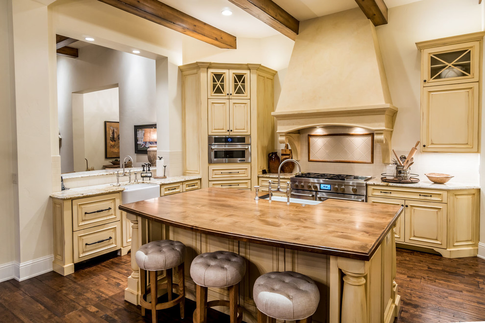 Photo of a large country l-shaped kitchen in Other with a farmhouse sink, raised-panel cabinets, beige cabinets, wood benchtops, beige splashback, ceramic splashback, panelled appliances, dark hardwood floors, with island and brown floor.