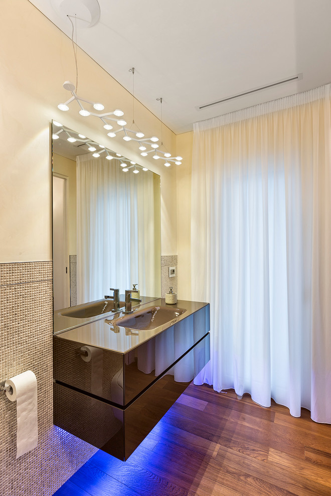 Mid-sized contemporary master bathroom in Bari with flat-panel cabinets, brown cabinets, a drop-in tub, a shower/bathtub combo, a two-piece toilet, beige tile, mosaic tile, beige walls, medium hardwood floors, an integrated sink and glass benchtops.