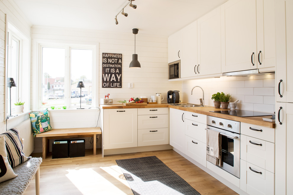 This is an example of a mid-sized transitional l-shaped kitchen in Malmo with a drop-in sink, shaker cabinets, white cabinets, wood benchtops, white splashback, subway tile splashback and medium hardwood floors.