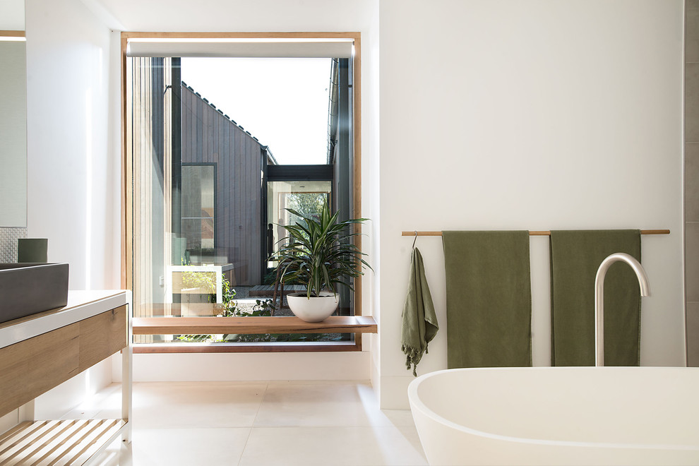 Photo of a large contemporary kids bathroom in Melbourne with furniture-like cabinets, light wood cabinets, a freestanding tub, an alcove shower, beige tile, porcelain tile, white walls, porcelain floors, an undermount sink, solid surface benchtops, beige floor, an open shower and white benchtops.