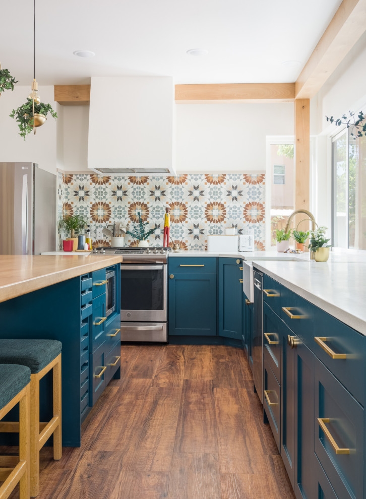 Mid-sized eclectic l-shaped eat-in kitchen in Other with a farmhouse sink, shaker cabinets, blue cabinets, wood benchtops, multi-coloured splashback, ceramic splashback, stainless steel appliances, medium hardwood floors, with island, white benchtop and exposed beam.