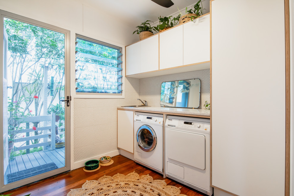 Mid-sized midcentury single-wall laundry room in Sunshine Coast with a drop-in sink, white cabinets, terrazzo benchtops, white splashback, mosaic tile splashback, white walls, dark hardwood floors, a side-by-side washer and dryer, brown floor and multi-coloured benchtop.
