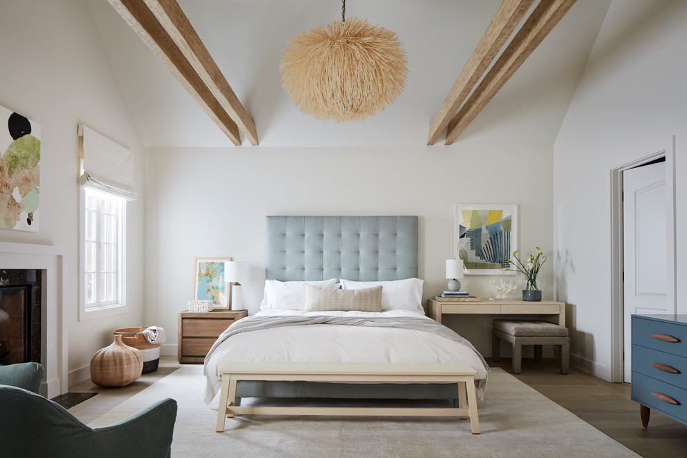 This is an example of a large beach style master bedroom in San Luis Obispo with white walls, a standard fireplace, medium hardwood floors, a wood fireplace surround and brown floor.