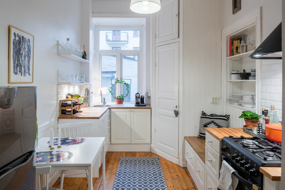 Photo of a scandinavian single-wall eat-in kitchen in Stockholm with a drop-in sink, raised-panel cabinets, white cabinets, wood benchtops, white splashback, subway tile splashback, black appliances, light hardwood floors and no island.