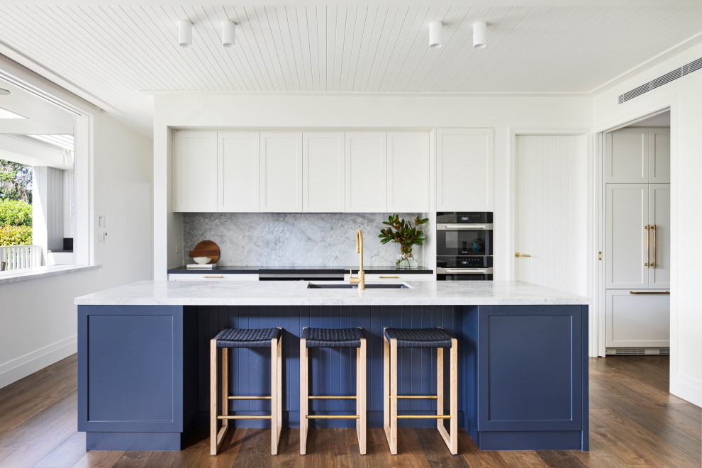 Inspiration for a mid-sized beach style open plan kitchen in Sydney with a drop-in sink, shaker cabinets, white cabinets, marble benchtops, grey splashback, marble splashback, black appliances, medium hardwood floors, with island, brown floor, grey benchtop and timber.