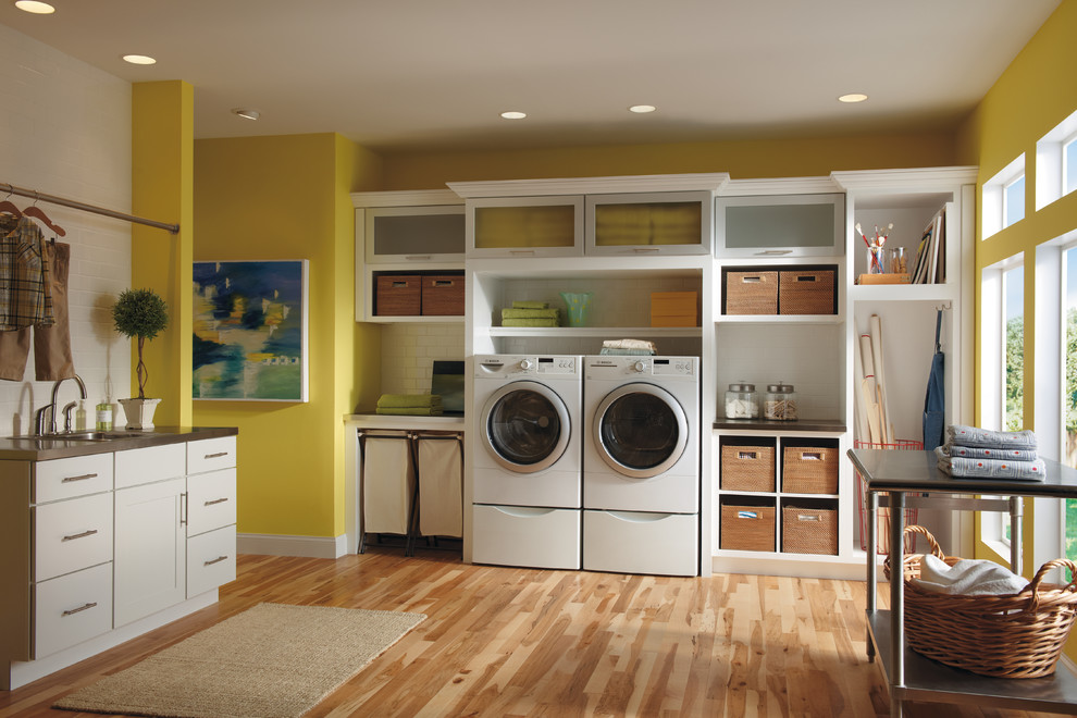 Photo of a large contemporary utility room in Other with a drop-in sink, shaker cabinets, white cabinets, yellow walls, light hardwood floors and a side-by-side washer and dryer.