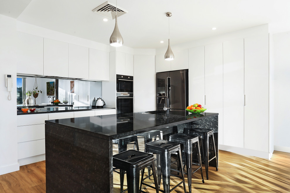 Idéer för mellanstora funkis svart l-kök, med en undermonterad diskho, vita skåp, granitbänkskiva, stänkskydd med metallisk yta, spegel som stänkskydd, svarta vitvaror, laminatgolv och brunt golv