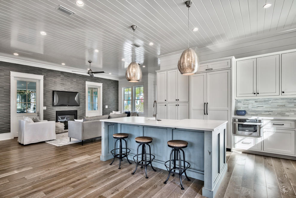This is an example of a mid-sized beach style u-shaped eat-in kitchen in Miami with a farmhouse sink, shaker cabinets, white cabinets, quartzite benchtops, grey splashback, stone tile splashback, stainless steel appliances, light hardwood floors, with island, brown floor and white benchtop.