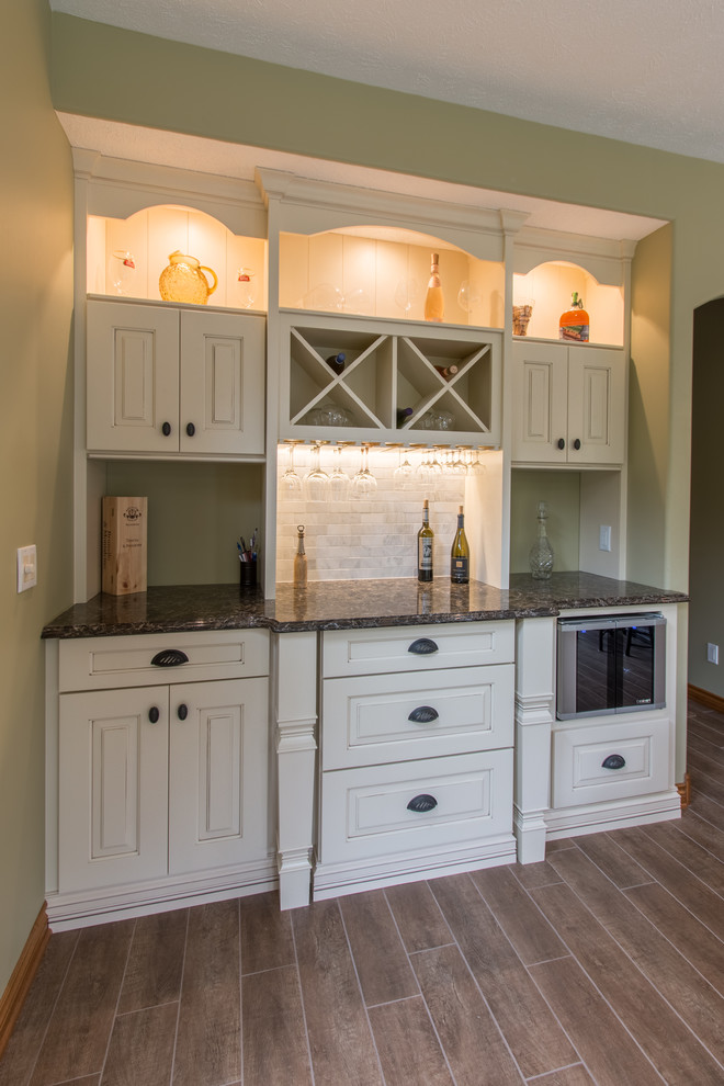 Small traditional single-wall wet bar in Grand Rapids with no sink, raised-panel cabinets, brown floor, white cabinets, granite benchtops, grey splashback, stone tile splashback and medium hardwood floors.