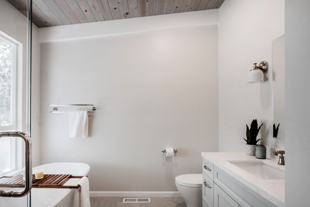 Идея дизайна: большая главная ванная комната в стиле неоклассика (современная классика) с фасадами с утопленной филенкой, синими фасадами, отдельно стоящей ванной, угловым душем, унитазом-моноблоком, бежевой плиткой, керамогранитной плиткой, бежевыми стенами, полом из керамогранита, врезной раковиной, столешницей из искусственного кварца, бежевым полом, душем с распашными дверями, белой столешницей, нишей, тумбой под две раковины, встроенной тумбой и деревянным потолком