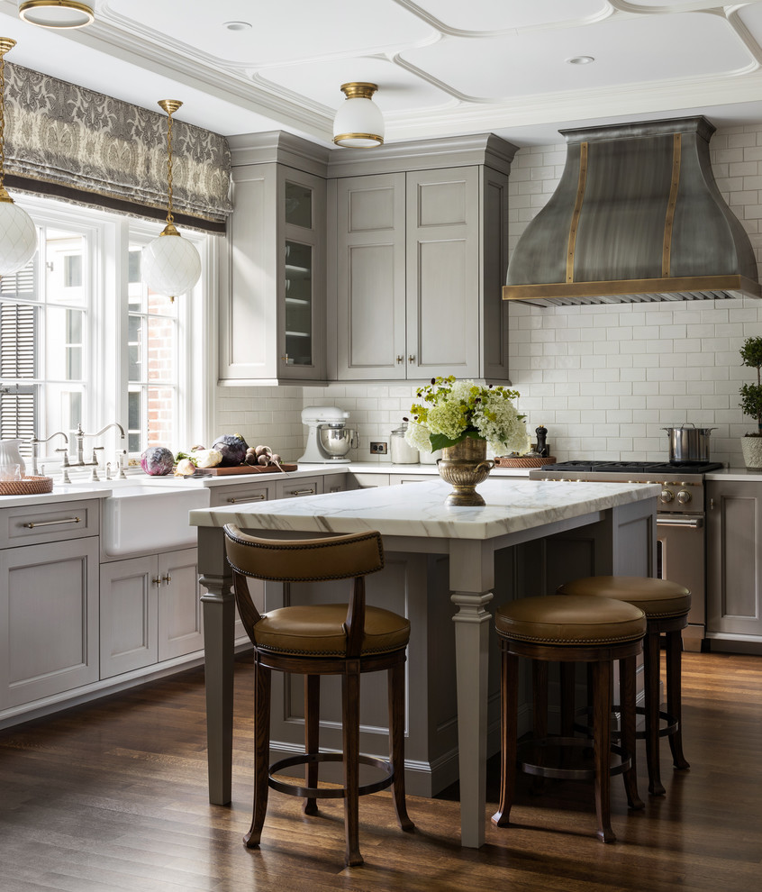 This is an example of a traditional l-shaped kitchen in New York with a farmhouse sink, recessed-panel cabinets, grey cabinets, white splashback, subway tile splashback, stainless steel appliances, dark hardwood floors, with island, brown floor and white benchtop.