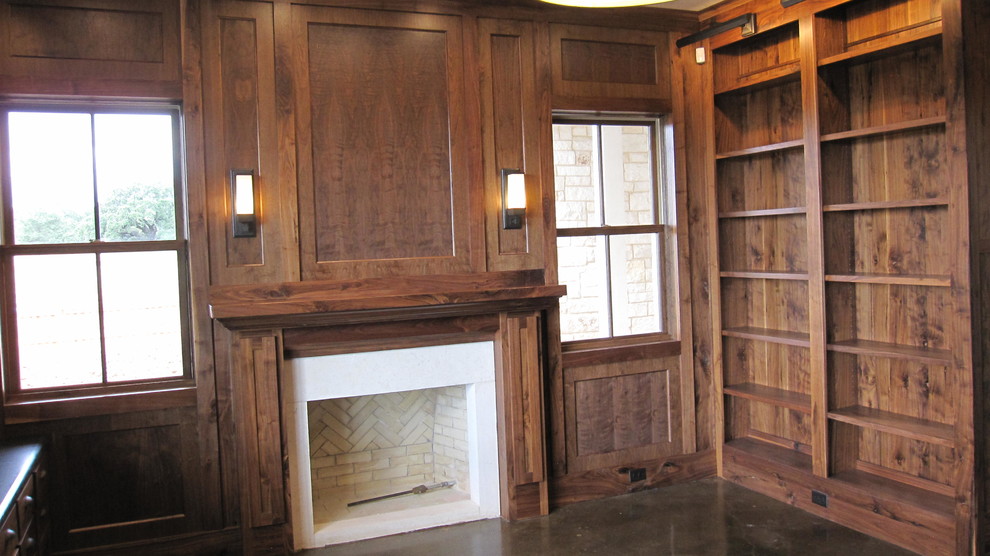 Photo of a large country home office in Austin with a library, brown walls, concrete floors, a standard fireplace, a brick fireplace surround, a built-in desk, brown floor, wood and wood walls.