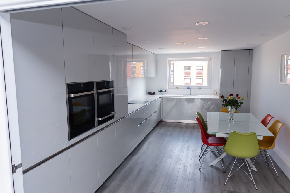 Mid-sized contemporary l-shaped eat-in kitchen in Other with a single-bowl sink, flat-panel cabinets, grey cabinets, solid surface benchtops, white splashback, black appliances, vinyl floors, no island, grey floor and white benchtop.
