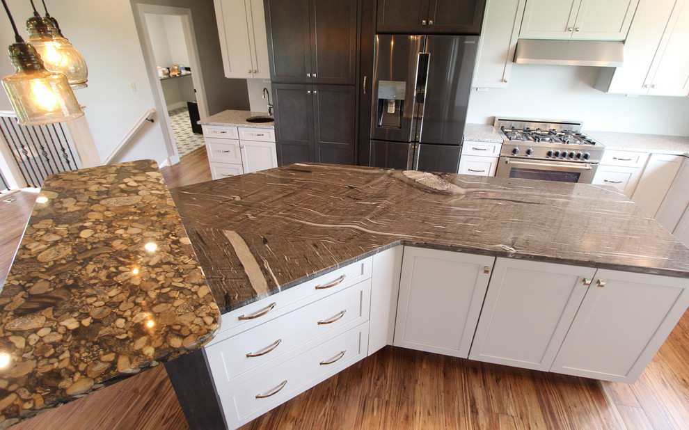 This is an example of a large transitional l-shaped kitchen in Cleveland with flat-panel cabinets, white cabinets, with island, a farmhouse sink, granite benchtops, white splashback, ceramic splashback, stainless steel appliances, medium hardwood floors, brown floor and multi-coloured benchtop.