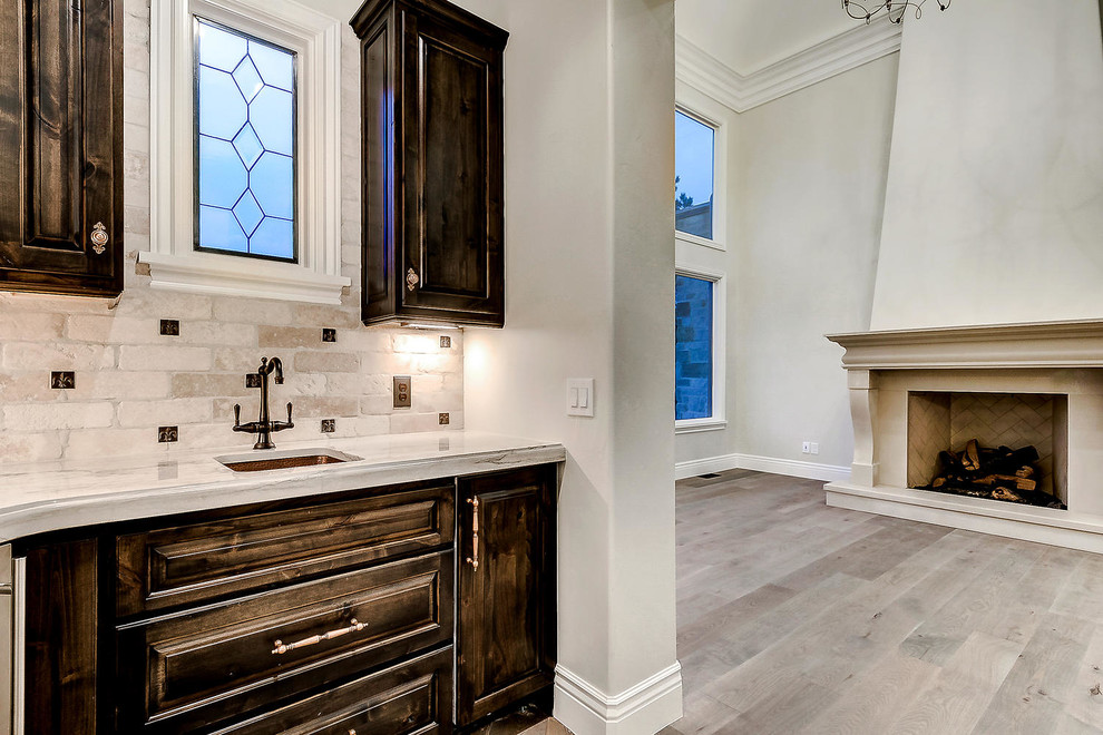 This is an example of a small traditional single-wall wet bar in Denver with an undermount sink, raised-panel cabinets, dark wood cabinets, marble benchtops, beige splashback, stone tile splashback, light hardwood floors, brown floor and beige benchtop.