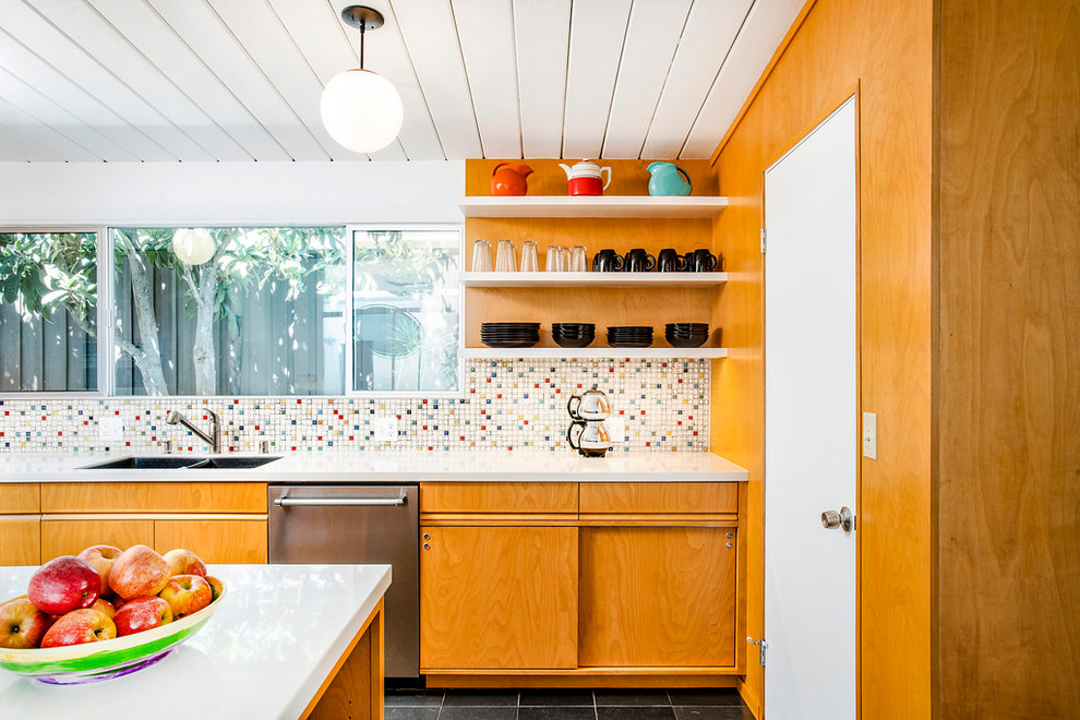 This is an example of a mid-sized midcentury galley open plan kitchen in Los Angeles with an undermount sink, flat-panel cabinets, multi-coloured splashback, stainless steel appliances, a peninsula, black floor, white benchtop, medium wood cabinets, solid surface benchtops, mosaic tile splashback and slate floors.