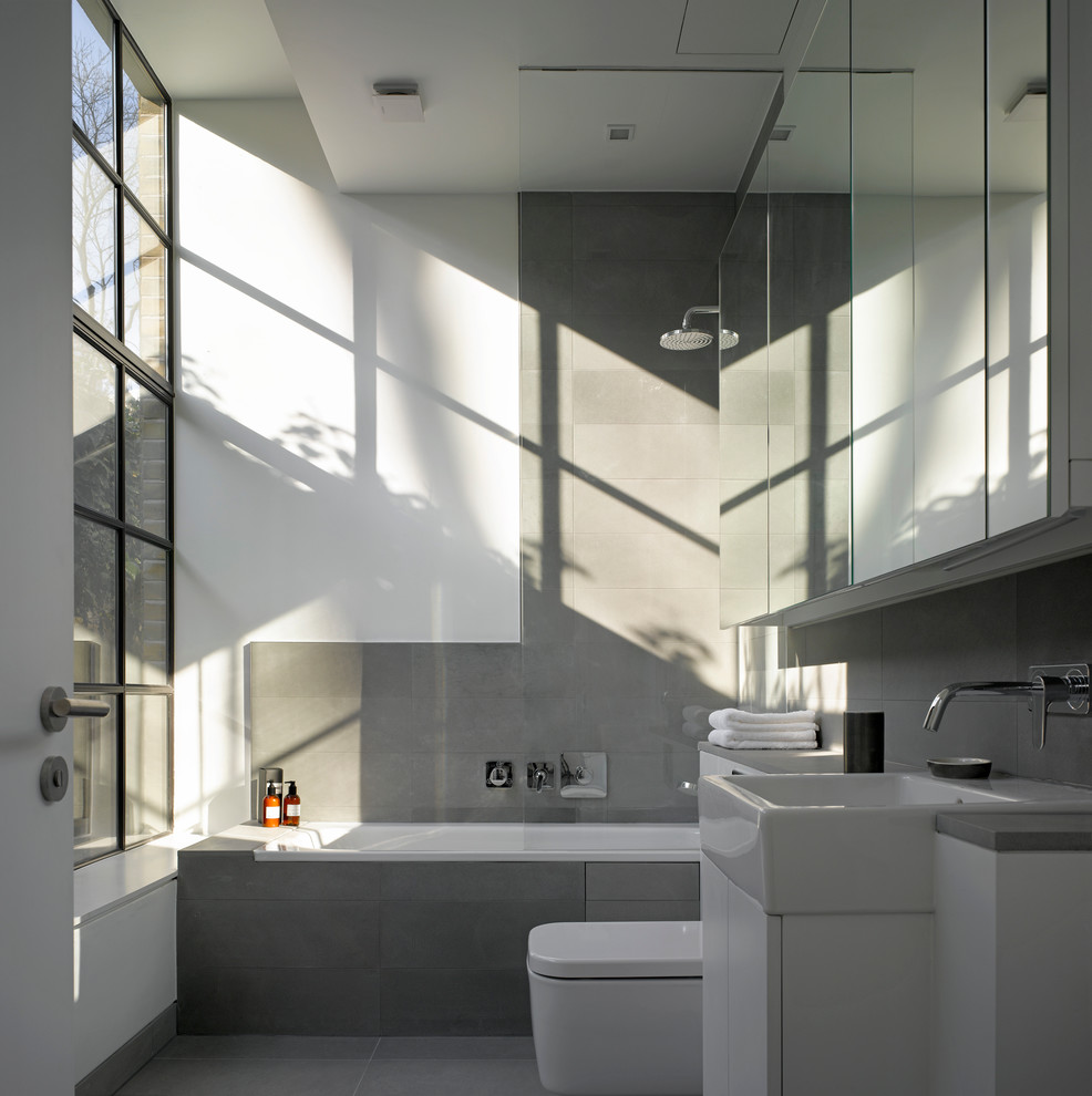 Photo of a modern bathroom in London with a vessel sink, white cabinets, a drop-in tub, a shower/bathtub combo, a wall-mount toilet and gray tile.