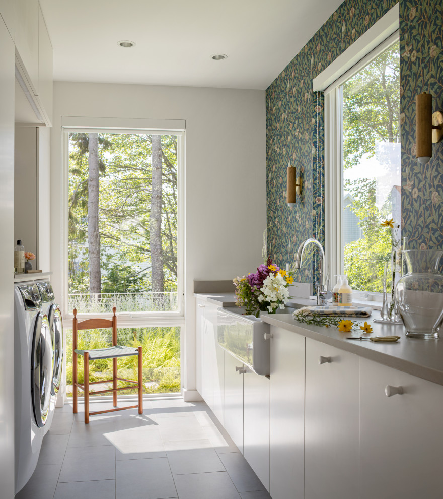 This is an example of a galley dedicated laundry room in Portland Maine with a farmhouse sink, flat-panel cabinets, white cabinets, grey walls, a side-by-side washer and dryer, grey floor, grey benchtop and wallpaper.
