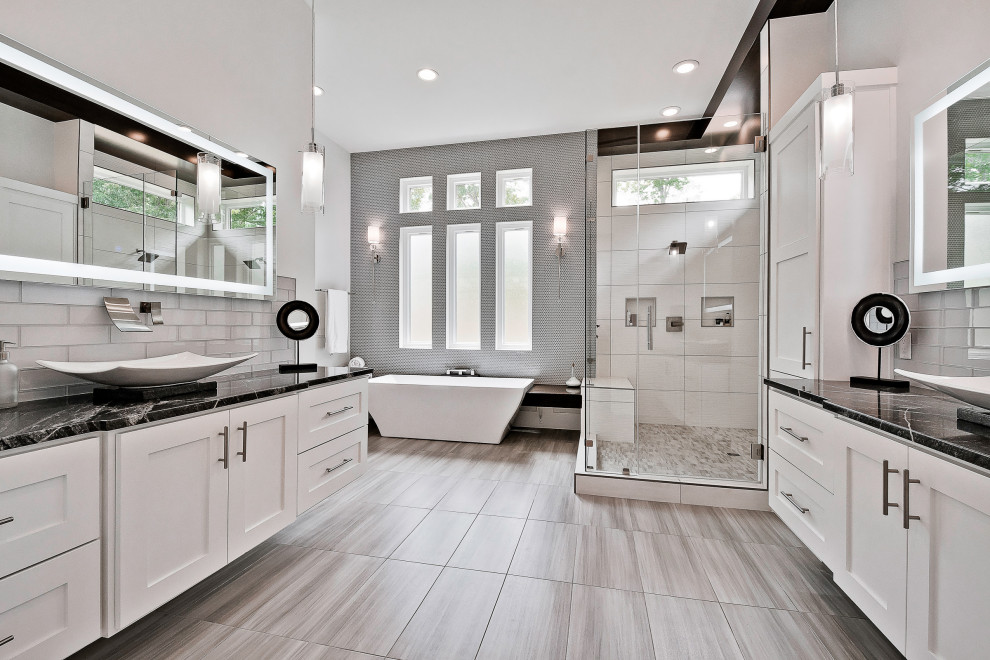 Photo of a large modern master bathroom in Other with recessed-panel cabinets, white cabinets, a freestanding tub, a double shower, a one-piece toilet, gray tile, glass tile, white walls, porcelain floors, a trough sink, quartzite benchtops, grey floor, a hinged shower door, black benchtops, a shower seat, a double vanity, a floating vanity and wood walls.