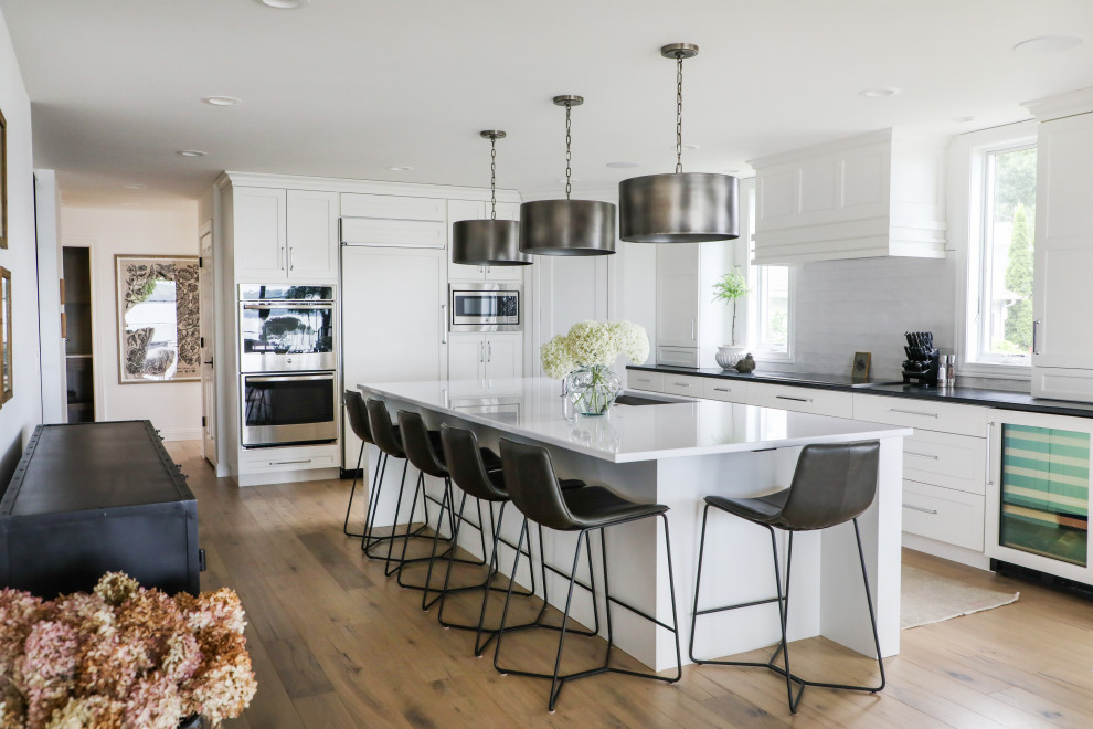 This is an example of a large transitional l-shaped eat-in kitchen in Other with an undermount sink, recessed-panel cabinets, white cabinets, quartz benchtops, white splashback, ceramic splashback, stainless steel appliances, medium hardwood floors, with island, brown floor and white benchtop.