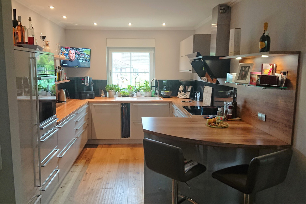 This is an example of a large contemporary u-shaped open plan kitchen in Berlin with a drop-in sink, flat-panel cabinets, grey cabinets, solid surface benchtops, black splashback, glass sheet splashback, black appliances, medium hardwood floors, no island, brown floor and brown benchtop.
