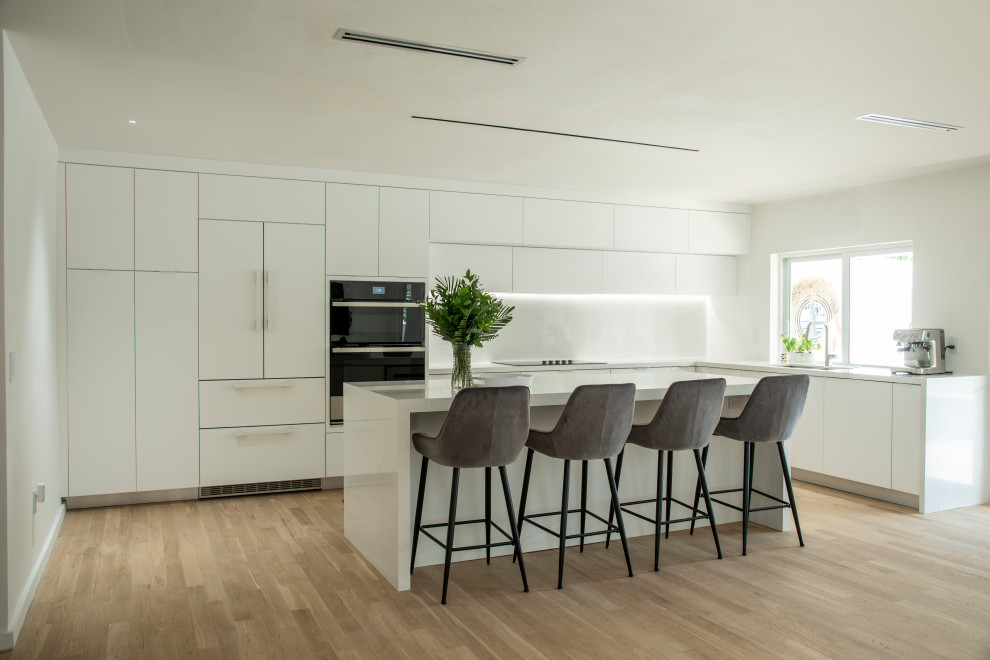 Large minimalist l-shaped medium tone wood floor and brown floor open concept kitchen photo in Miami with an undermount sink, flat-panel cabinets, white cabinets, quartz countertops, white backsplash, quartz backsplash, paneled appliances, an island and white countertops