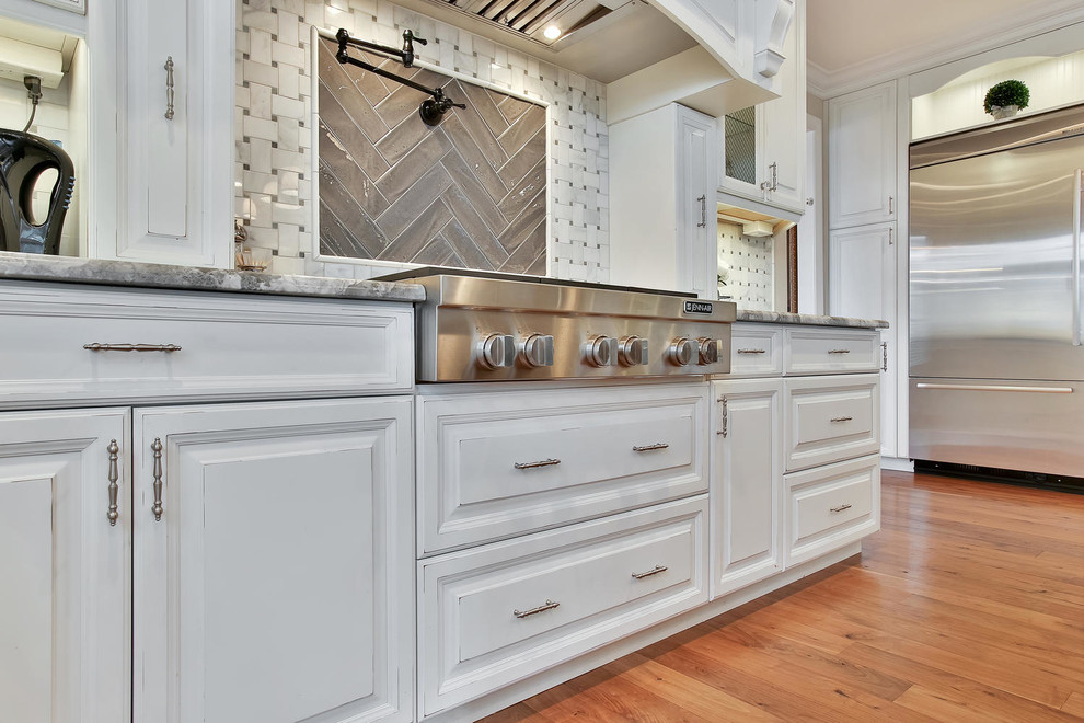 This is an example of a mid-sized beach style eat-in kitchen in New York with a farmhouse sink, raised-panel cabinets, distressed cabinets, granite benchtops, grey splashback, ceramic splashback, stainless steel appliances, medium hardwood floors and with island.