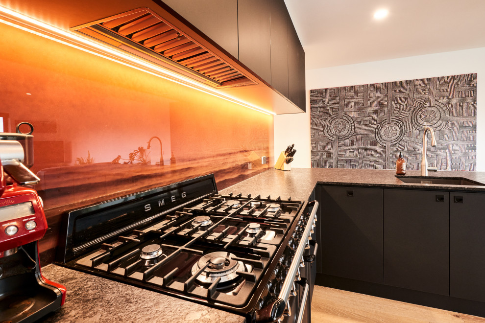 На фото: п-образная кухня-гостиная среднего размера в стиле модернизм с плоскими фасадами, черными фасадами, столешницей из кварцевого агломерата, оранжевым фартуком, фартуком из стекла, черной техникой и черной столешницей с