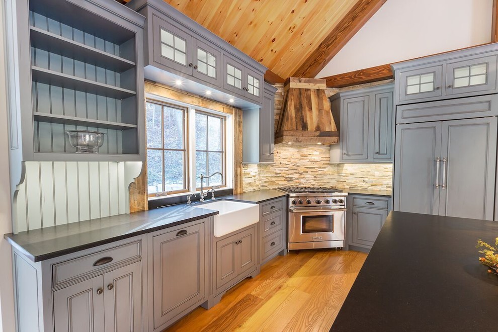Photo of a mid-sized country l-shaped open plan kitchen in Burlington with a farmhouse sink, beaded inset cabinets, grey cabinets, soapstone benchtops, stone tile splashback, stainless steel appliances, light hardwood floors and with island.