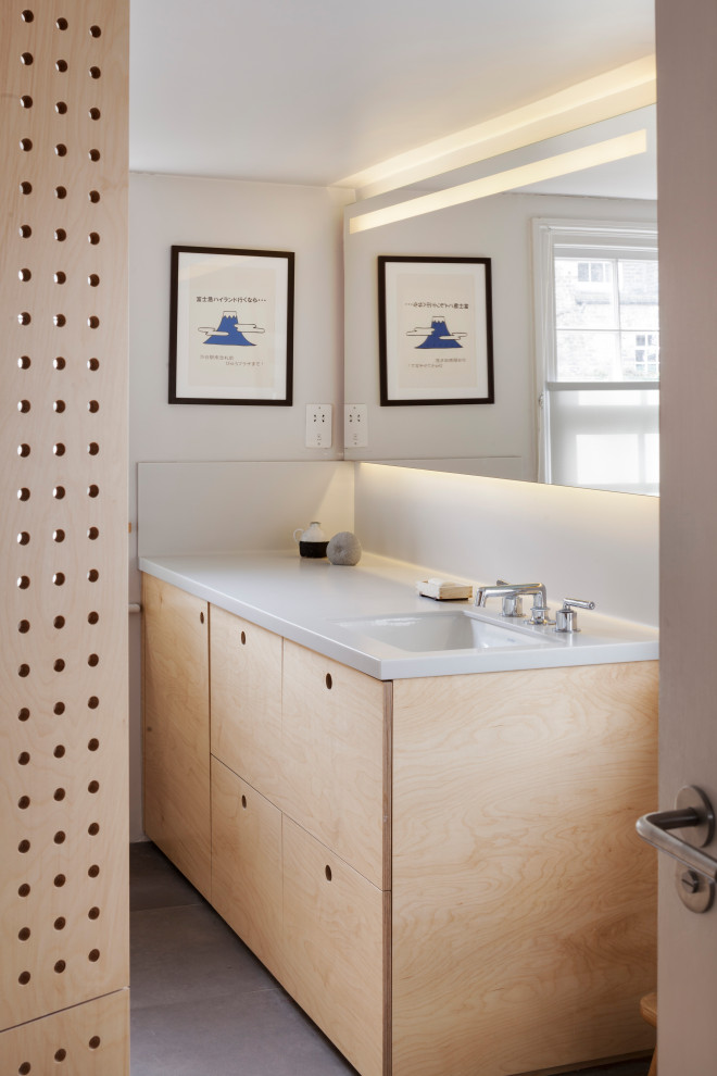 This is an example of a small contemporary kids bathroom in London with flat-panel cabinets, light wood cabinets, a drop-in tub, a shower/bathtub combo, a wall-mount toilet, gray tile, ceramic tile, grey walls, ceramic floors, a drop-in sink, solid surface benchtops, grey floor, a shower curtain, grey benchtops, a laundry, a single vanity, a built-in vanity and panelled walls.